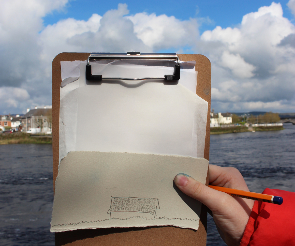 Childs hand holding up clipboard with drawing of a house on it. Background is a rive and some boats